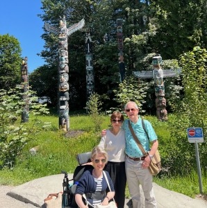 A woman sitting in a wheelchair beside her family with the Stanley Park Totem Poles in the background.